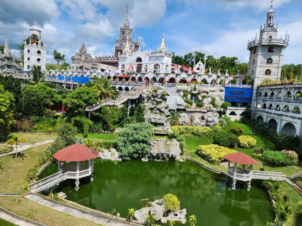 Simala Shrine 2024: A Journey to the Miraculous and Mystical Castle Church in Cebu 1 It is a picture of Simala Shrine in Cebu There is a castle looking church with a huge flower garden and a pool with two cottages above the pool.