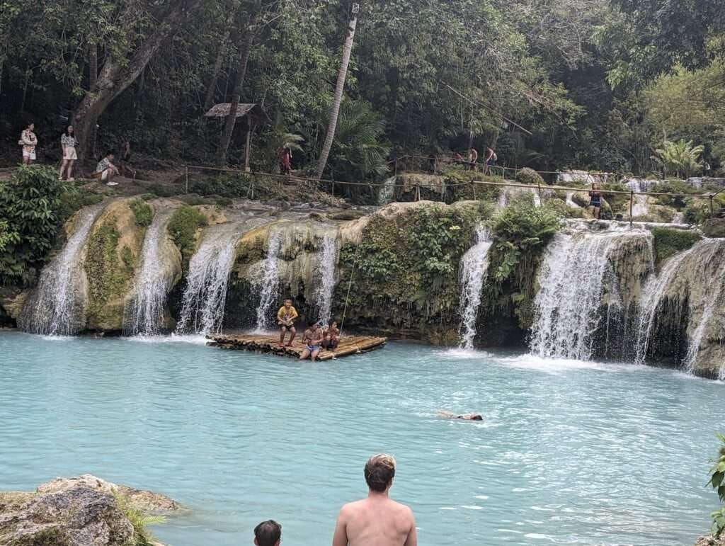 Cambugahay Falls: Complete Siquijor Island Waterfall Guide 5 People swimming, jumping off cliffs, and relaxing by the turquoise water at Cambugahay Falls. Lush greenery surrounds the cascading waterfalls